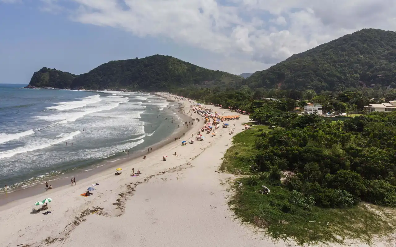 Vista panorâmica da Praia de Camburi em São Sebastião, SP. Faixa de areia clara com mar azul e morros cobertos por Mata Atlântica exuberante ao fundo.