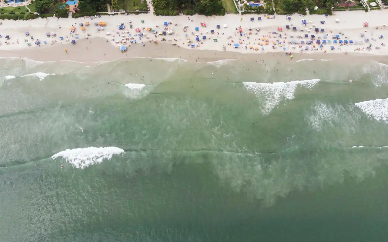Vista aérea da Praia de Juquehy em São Sebastião, SP. Extensa faixa de areia clara com mar calmo e esverdeado, ideal para famílias, cercada pela Mata Atlântica e infraestrutura costeira.